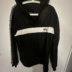 Svart vindjacka från Alpha Industries - Snygg svart vindjacka från Alpha Industries med vit logga och vit rand över bröstet. Jackan har huva och cool text längs ärmarna. Perfekt för dig som gillar streetwear och vill ha en jacka med attityd.