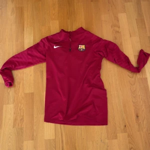 Vinröd Nike Barcelona half zip tröja - Säljer en vinröd långärmad träningströja från Nike med FC Barcelona-logga på bröstet. Tröjan har en half zip-dragkedja vid halsen och är perfekt för fotbollsträning eller chill. Snygg och sportig design!