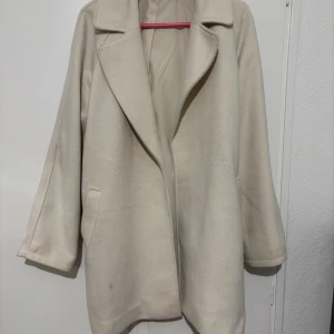 Beige kappa med bälte och krage - Stilren beige kappa med klassisk krage och bälte i midjan. Jackan har raka ärmar, två fickor framtill och en mjuk, slät yta. Perfekt för dig som gillar en clean och tidlös look under kyliga dagar.