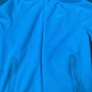 Blå Nike NOCTA vindjacka med huva - Säljer en blå vindjacka från Nike NOCTA med huva och dragkedja framtill. Jackan har två fickor med dragkedja och en stilren look med diskret Nike-logga på bröstet. Perfekt för dig som gillar sportig och clean stil. Använd cirka 4 gånger inte mer storlek L 