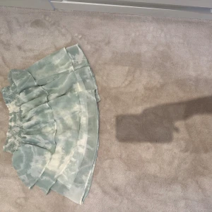 Grönvit volangkjol från H&M - Supersöt kort volangkjol från H&M i ljusgrönt och vitt med batikmönster. Kjolen har resår i midjan och flera lager för extra volym. Perfekt för dig som gillar en luftig och trendig stil.