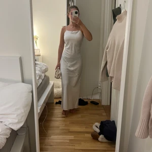 Vit långklänning med smala axelband - Säljer en stilren vit långklänning med smala axelband och rak passform. Klänningen har en minimalistisk look och knytning på axlarna. Perfekt för dig som gillar enkel och elegant stil. Materialet känns lätt och svalt, perfekt för varmare dagar. Använd 1 gång 