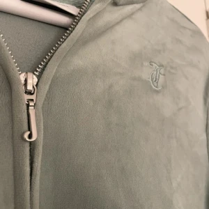 Grön hoddie från Juicy Couture - Mjuk och lyxig ljusgrön hoodie från Juicy Couture i velour med dragkedja framtill, huva och broderad JC-logga på bröstet. Klassisk känguruficka och ribbade muddar. Perfekt för en chill och trendig look.
