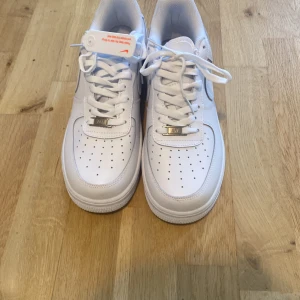 Nike Air Force 1 vita sneakers - Klassiska Nike Air Force 1 sneakers i helvitt skinn med perforerad tå, snörning och metall-detalj vid snörningen. Snygg och tidlös modell med rund tå och platt sula. Perfekta för dig som vill ha en clean och fräsch look.