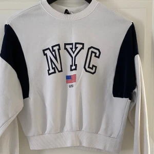 Vit och marinblå NYC sweatshirt Divided - Säljer en vit sweatshirt från Divided med marinblå detaljer på axlarna och ärmarna. Framsidan har tryck med texten 'NYC' och en liten amerikansk flagga. Tröjan har rund hals, ribbade muddar och är i mjuk bomull. Perfekt för en chill och sportig look.