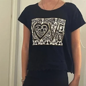 Svart topp med LOVE-tryck och spets - Svart topp med korta ärmar och croppad passform. Framsidan har ett stort vitt tryck med svart spetsmönster och texten LOVE där O:et är format som ett hjärta. Materialet känns mjukt och toppen har en avslappnad look.