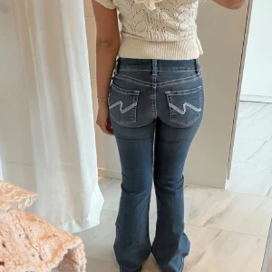 Blå bootcut jeans med broderade fickor - Snygga blå jeans med bootcut-modell och låg midja. Jeansen har vita broderade detaljer på bakfickorna och klassisk femficksdesign. Materialet är jeans och passformen är normal med lätt utsvängda ben.