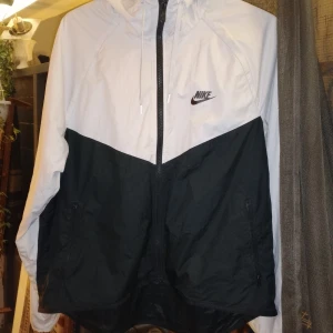 Nike svartvit vindjacka med huva - Nike vindjacka i svart och vitt med snygg tvåfärgad design. Jackan har huva med snörning, hel dragkedja och två sidofickor med dragkedja. Perfekt för dig som gillar sportig stil och vill ha något lätt och funktionellt.