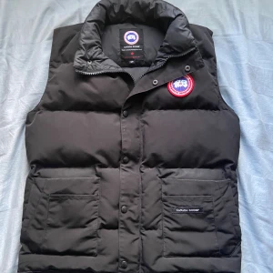 Svart dunväst från Canada Goose - Svart dunväst från Canada Goose med hög krage, två stora fickor framtill och klassisk logotyp på bröstet. Västen har tryckknappar och dragkedja framtill. Perfekt för lager-på-lager och riktigt snygg till streetwear.