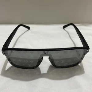 Louis Vuitton glasögon  - Säljer ett par svarta solglasögon från Louis Vuitton med stora, rektangulära glas och tydligt LV-monogram över hela linsen. Bågarna är i svart plast och har en modern, kantig design som sticker ut.