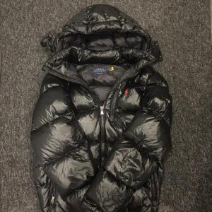 Svart pufferjacka från Polo Ralph Lauren - Säljer en svart pufferjacka från Polo Ralph Lauren med huva och dragkedja. Jackan är i extremt bra skick och endast använd fåtal gånger. Pris ej hugget i sten