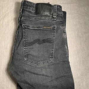 Svarta jeans från Nudie Jeans Co, 27/32 - Snygga svarta jeans från Nudie Jeans Co med klassisk femficksdesign och coola kontrastsömmar. Jeansen har en mörk tvätt och subtilt broderad bakficka. Passar dig som gillar stilrena och tidlösa jeans med en modern touch.