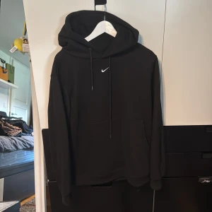 Nike hoodie - Svart hoodie från Nike med klassisk vit swoosh på bröstet. Tröjan har huva med snörning, stor känguruficka och ribbade muddar. Mjuk fleece på insidan gör den extra skön. Perfekt för chill eller träning. Aldrig använd.
