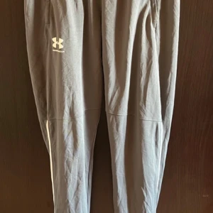 Grå träningsbyxor från Under Armour - Snygga grå mjukisbyxor från Under Armour med logga på låret. Byxorna har en slim passform och är gjorda i ett mjukt och stretchigt syntetmaterial. Perfekta för träning eller chill. Elastisk midja och smalare benavslut.