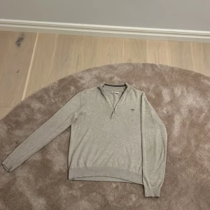 Beige half zip tröja från GANT - Snygg beige långärmad tröja från GANT med half zip och liten broderad logga på bröstet. Tröjan har ribbad krage och muddar, perfekt för dig som gillar stilren och klassisk look. Materialet är mjukt och känns skönt mot huden.