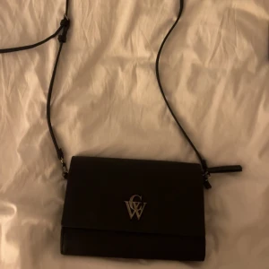 Svart axelväska från CW - Snygg svart axelväska från CW med stilren design och silverfärgat logospänne framtill. Väskan har justerbar axelrem, magnetlås och dragkedja inuti. Tillverkad i slätt skinn och har flera fack för smart förvaring. Perfekt storlek för det viktigaste. Lite sliten men går såklart att rengöra, inga andra fel😇Priset är 500kr om man köper en helt ny! 