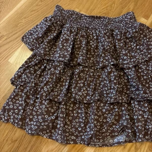 Brun blommig volangkjol - Supersöt brun kortkjol med små vita blommor och flera volanger. Kjolen har resår i midjan för en bekväm passform och är gjord i ett lätt och luftigt tyg, perfekt för varmare dagar. Den har en flowy känsla och passar perfekt till en enkel topp.