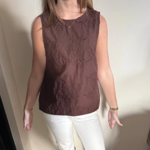 Brun ärmlös blus med broderade blommor - Säljer en brun ärmlös blus med stora broderade blommor. Toppen har rak passform och rund halsringning, perfekt för dig som gillar en stilren men ändå unik look. Materialet känns mjukt och luftigt, och de broderade detaljerna ger en extra touch.
