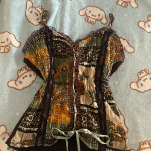 Vintage Xanaka Boho-inspirerad topp med snöre  - Säljer en boho-inspirerad korsettopp med smala axelband, snörning framtill och volangkant vid bysten. Toppen har ett patchwork-liknande mönster i grönt, brunt, beige och orange med svarta spetsdetaljer. Materialet är mjukt och glansigt framtill, baksidan är brun och stretchig. Toppen är från Xanaka och är gjort i Frankrike. Skickar snabbt köp 🙂‍↕️
