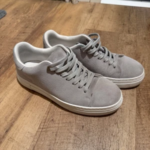 Grå mockasneakers från Selected Homme - Snygga grå sneakers från Selected Homme i mocka med klassisk låg profil och vita sulor. Skorna har rund tå, snörning och stilren design som passar till det mesta. Perfekta för dig som gillar en clean och modern look. Skorna är nästan nya använt dem cirka 2 gånger, nypris är 1200 kr säljer för 800 kr