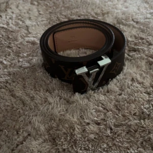 bälte - Snyggt bälte i brunt skinn med klassiskt monogrammönster och silverfärgat spänne. Bältet har beige insida och tydliga logodetaljer. Perfekt accessoar för att lyfta din outfit med en lyxig touch.