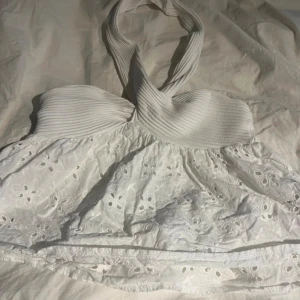 Vit halterneck topp med broderi - Supersöt vit halterneck topp med ribbad överdel och broderade blommor på den vida nederdelen. Toppen har omlottkrage och är ärmlös, nästintill nyskick har använts 1-2 gånger