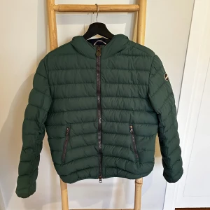 Grön pufferjacka från Colmar - Säljer en mörkgrön pufferjacka från Colmar med horisontella sömmar och två dragkedjeförsedda fickor framtill. Jackan har en hög krage och klassisk Colmar-logga på ärmen. Perfekt för kyliga dagar och riktigt snygg till streetwear. Jackan är lagad professionellt på ena ärmen vilket syns på sista bilden. Storlek 46 är en stor small eller liten medium.