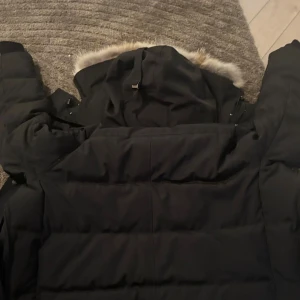 Svart dunjacka från Canada Goose - Säljer en svart dunjacka från Canada Goose med klassisk patch på ärmen. Jackan har huva med fuskpäls, dragkedja och knappar framtill samt två stora fickor. Perfekt för kalla vinterdagar och riktigt snygg streetstil.