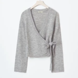 Grå omlottkofta från Gina tricot  - Supermjuk grå omlottkofta från Gina tricot med v-ringning och knyt i sidan. Koftan är stickad, har lång ärm och croppad passform. Perfekt att slänga över ett linne eller t-shirt för en chill look.