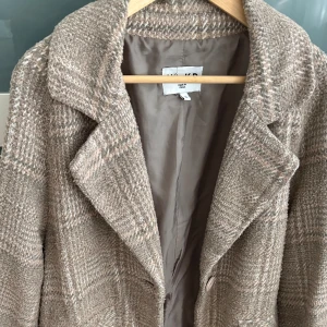 Beige rutig kappa från NAKD - Säljer en beige rutig kappa från Zara med klassisk krage och knappar framtill. Kappan har en snygg oversized passform och är tillverkad i ett ullblandat material som ger en mjuk och varm känsla. Perfekt för dig som gillar stilrena och trendiga ytterplagg.