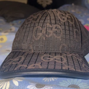 Svart keps med GG-mönster - Säljer en svart keps med klassiskt GG-mönster och rutigt print över hela kepsen. Kepsen har böjd skärm och är tillverkad i ett tygmaterial med diskret glans. Perfekt accessoar för en trendig look.