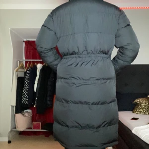 Svart lång pufferjacka med fickor - Säljer en svart lång pufferjacka med hög krage och stora fickor framtill. Jackan har elastisk midja och är riktigt tjock och varm, perfekt för kalla dagar. Stängs med dragkedja och har ribbade muddar vid ärmslut. Snygg och praktisk för vintern!