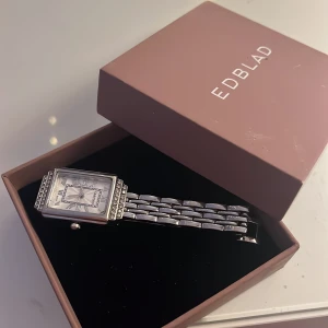 Silverfärgad klocka  - Stilren klocka i silverfärg med rektangulär urtavla och romerska siffror. Klockan har ett armband i metall med snygga länkar och dekorativa stenar runt urtavlan. Perfekt accessoar för att lyfta din outfit.