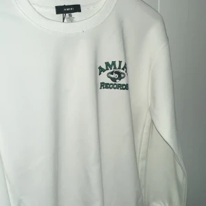 Vit amiri sweatshirt  - Vit sweatshirt från Amiri med grönt och svart Amiri Records-tryck fram och stort tryck på ryggen. Klassisk rund halsringning och långa ärmar. Skön och stilren tröja i bomull, perfekt för dig som gillar streetwear med coola detaljer.