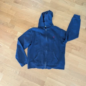 Mörkblå hoodie från Polo Ralph Lauren strl 164 - Mörkblå hoodie från Polo Ralph Lauren i storlek XL (18-20) storlek stämmer inte alls i mina ögon!! Den här tröjan motsvarar en killstorlek 164 (11-14 år) jag är 173 cm lång och tröjan är för kort i armarna på mig...Klassisk design med dragkedja, huva och kängurufickor. Broderad röd logga på bröstet. Tillverkad i mjuk bomullsmix, perfekt för chill dagar eller när du vill ha en enkel streetlook. En litet hål i vänster arm på baksidan. Se bild. 