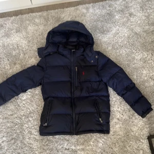 Mörkblå dunjacka från Polo Ralph Lauren - Säljer en mörkblå dunjacka från Polo Ralph Lauren med huva och dragkedja. Jackan har två sidofickor med dragkedja och ett rött Polo-logomärke på bröstet. Perfekt för kalla dagar och har en schysst, puffig look.