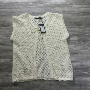 Vit stickad bolerotopp från 157 - Supersöt vit stickad bolerotopp från 157 med knyt framtill. Toppen har ett genomgående hålmönster, volangkant vid ärmslut och nederkant samt v-ringning. Perfekt att slänga över ett linne eller klänning för en extra touch.