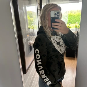 Camouflage hoodie från Converse - Säljer en grön camouflage hoodie från Converse med stor vit logga på bröstet och tryckt text på ärmen. Tröjan har huva och magficka, samt ribbade muddar vid ärmslut och nederkant. Perfekt för dig som gillar streetstyle och vill sticka ut.