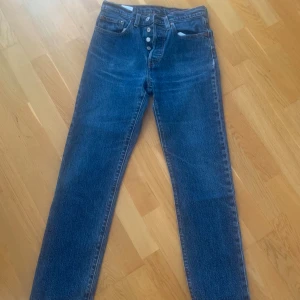 Levi's 501 blå jeans W27 L30 - Klassiska Levi's 501 jeans i blå denim med raka ben och knappgylf. Modellen är midjehög och har fem fickor samt Levi's Premium-märke. Perfekta för dig som gillar tidlös stil och schysst passform.
