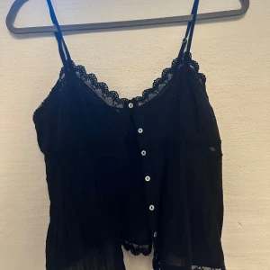Svart linnetopp med spets och knappar - Säljer en svart linnetopp med tunna axelband och dekorativ spets längs halsringning och nederkant. Toppen har vita knappar framtill och är lätt transparent med en luftig passform. Perfekt för dig som gillar en chill och feminin stil.