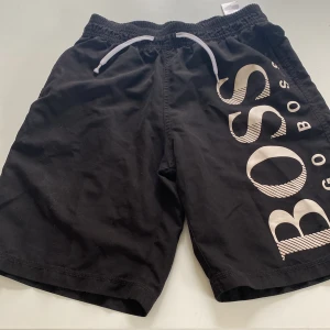 Badbyxor Hugo boss  - Svarta badbyxor från BOSS med vit snörning i midjan och stort BOSS-tryck längs ena benet. Sköna och avslappnade med elastisk midja, perfekta för sommardagar eller chill hemma. Klassisk design med modern touch. Snabb affär prioriteras. Mvh Anton