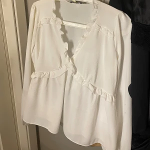 Vit blus med volang och v-ringning - Säljer en söt vit blus med v-ringning och volangdetaljer längs halsen och över bröstet. Blusen har långa ärmar och är tillverkad i ett lätt, mjukt tyg som ger ett flowy intryck. Perfekt för dig som gillar romantiska och feminina plagg.