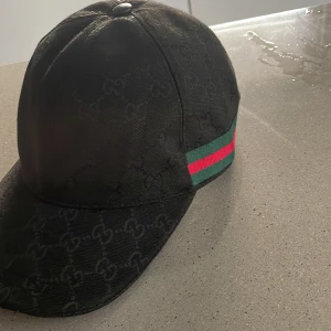 Svart Gucci keps med GG-mönster - Snygg svart keps från Gucci med diskret GG-monogram över hela ytan. Klassisk böjd skärm och justerbar rem baktill i skinn. Sidan har den ikoniska gröna och röda randen. Tillverkad i Italien, materialet är bomull och skinn. Perfekt för dig som vill ha en lyxig streetstyle. Pris går att diskutera 