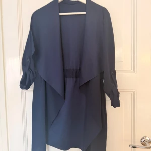 Mörkblå tunn trenchcoat - Snygg mörkblå tunn trenchcoat med långa ärmar som kan rullas upp och fästas med knapp. Jackan har en avslappnad passform och ett elastiskt parti i midjan för extra komfort. Perfekt för lager-på-lager och stilren look. Det står ingen storlek men jag har vanligtvis storlek S, så skulle säga att det är någonstans där. 