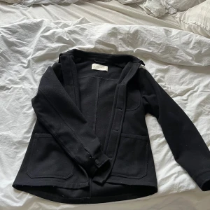 Svart overshirt från A Day's March - Stilren svart overshirt från A Day's March med klassisk krage, knäppning framtill och tre praktiska fickor. Jackan är tillverkad i mjuk bomull och har en rak passform som funkar till många olika outfits.