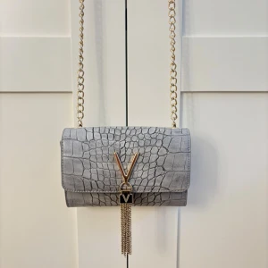Valentino väska - ”Valentino Bags DIVINA” Snygg grå axelväska från Valentino med krokopräglat mönster och guldfärgad kedja. Väskan har ett stort V i metall framtill och dekorativa guldkedjor som hänger ner. Perfekt storlek för det viktigaste och ger en lyxig vibe till din outfit. Hör med än gärna av dig vid frågor/ intresse! *NYSICK ALDRIG ANVÄNT *