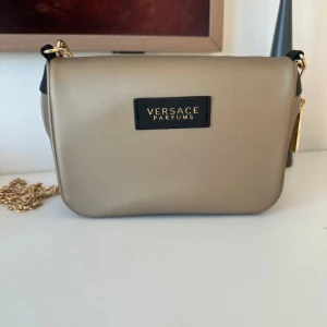 Beige axelväska från Versace Parfums - Snygg beige axelväska från Versace Parfums med guldfärgad kedja och dragkedja. Väskan har en stilren, rektangulär form och svart innerfoder med Versace-logga. Perfekt storlek för det viktigaste och har exklusiva detaljer i guld.