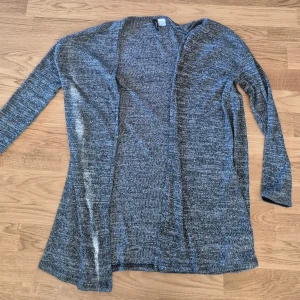 Grå lång kofta i stickat material - Mysig och lång grå kofta i stickat material. Koftan är öppen framtill och har långa ärmar. Perfekt att slänga på sig över en t-shirt eller topp för en avslappnad look.