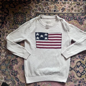 Beige Selected Homme tröja med USA-flagga – stl S, mycket bra skick - Säljer en snygg och stilren beige tröja från Selected Homme i storlek S. Tröjan har en stor USA-flagga framtill som ger den en sportig och preppy känsla. Tillverkad i mjukt material som sitter skönt mot kroppen, med klassiska detaljer som rund hals, ribbade muddar och avslappnad passform.  Plagget är i mycket bra skick, väl omhändertaget och utan några skador eller fläckar. Perfekt att bära både till vardags med jeans och sneakers eller som en del av en mer trendig lager-på-lager-look.