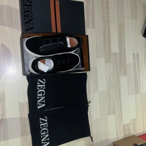 Svarta sneakers från Zegna, strl 44 - Snygga svarta sneakers från Zegna i storlek 44. Skorna har en stilren design med ovandel i kornigt skinn och detaljer i mocka, samt vit platt sula. Kommer med originalkartong och två svarta dustbags med Zegna-logga. Tveka inte med frågor eller bud 